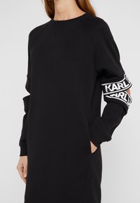 Donna che indossa un vestito felpa nero a maniche lunghe con maniche a taglio e bande con logo bianco sulle braccia superiori.