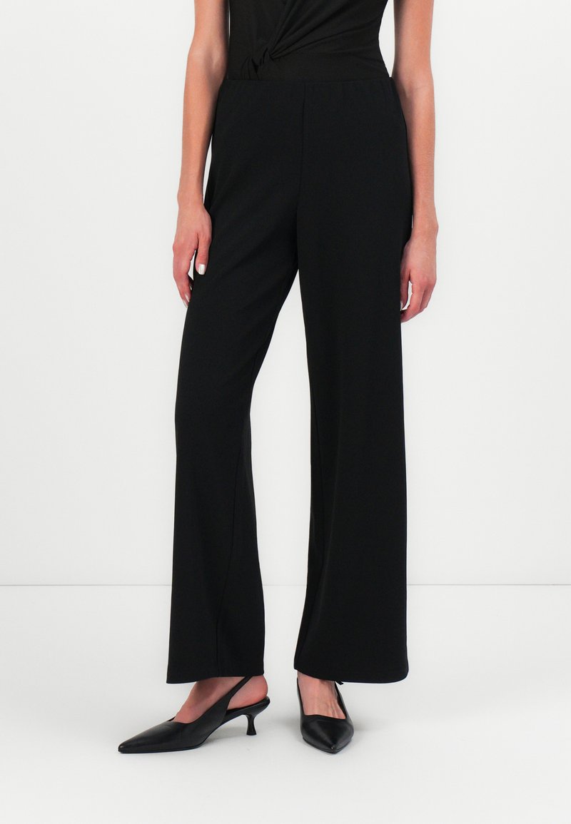 Schwarze weit geschnittene Hosen aus glattem Stoff mit elastischem Bund. Getragen mit spitzen schwarzen High Heels. Einfaches, modernes Design.