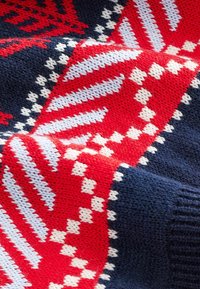 Gestrickter Pullover mit marineblauem Grundton und roten sowie weißen geometrischen Mustern, darunter Zickzacklinien und Rauten. Weiche Textur mit dickem Garn.