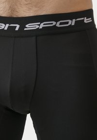 Shorts de sport noirs avec une texture lisse, dotés d'une large taille avec "sport" imprimé en blanc. Le tissu est extensible et ajusté.