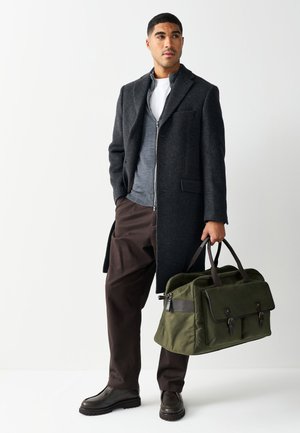 Mann trägt dunkelgrauen Mantel, grauen Reißverschluss-Pullover, braune Hose, braune Schuhe, hält grüne Tasche mit zwei Griffen vor weißem Hintergrund.