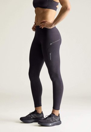 Personne en forme portant des leggings noirs taille haute et des baskets assorties avec poche zippée, debout les mains sur les hanches devant un fond uni.