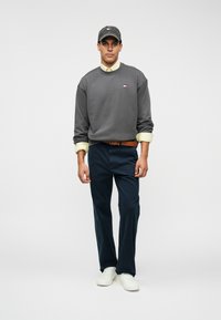 Γκρίζα φούτερ με στρογγυλή λαιμόκοψη, ναυτικές παντελόνες και λευκά αθλητικά παπούτσια. Περιλαμβάνει γκρι καπέλο με λογότυπο και κίτρινες μανσέτες που φαίνονται.