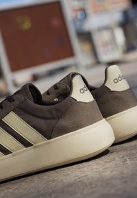 Een paar bruine en beige Adidas sneakers met witte zolen geplaatst op buitenverharding, met zichtbare zijstrepen en gebrande hiellabels.