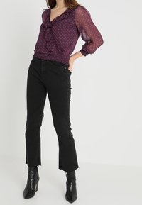 Lila transparente Bluse mit Rüschenkragen und weißen Pünktchen, kombiniert mit schwarzen hochgeschnittenen Jeans im ausgestellten Schnitt und fransigem Saum.