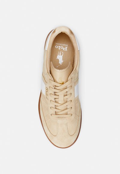 Beige suede sneaker with white side stripes, flat laces, and a round toe. Features a logo on the tongue and a textured interior.