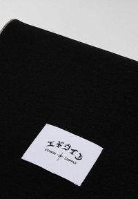 Tessuto nero con un'etichetta bianca con la scritta "エディウィン SUPPLY" e grafiche stilizzate. Il tessuto ha una texture liscia e opaca.