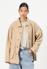 Beige oversized jacket with a shirt collar, front pockets, and button closure. Worn over a light gray t-shirt and loose denim jeans.