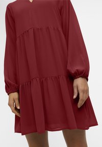 Robe rouge à volants avec manches longues, confectionnée dans un tissu léger. Elle présente un col en V et des poignets froncés, créant une silhouette fluide.