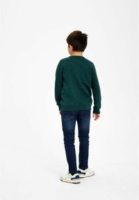 Niño de pie de espaldas, vistiendo un sudadera verde oscura, jeans azules y zapatillas blancas, contra un fondo blanco liso.