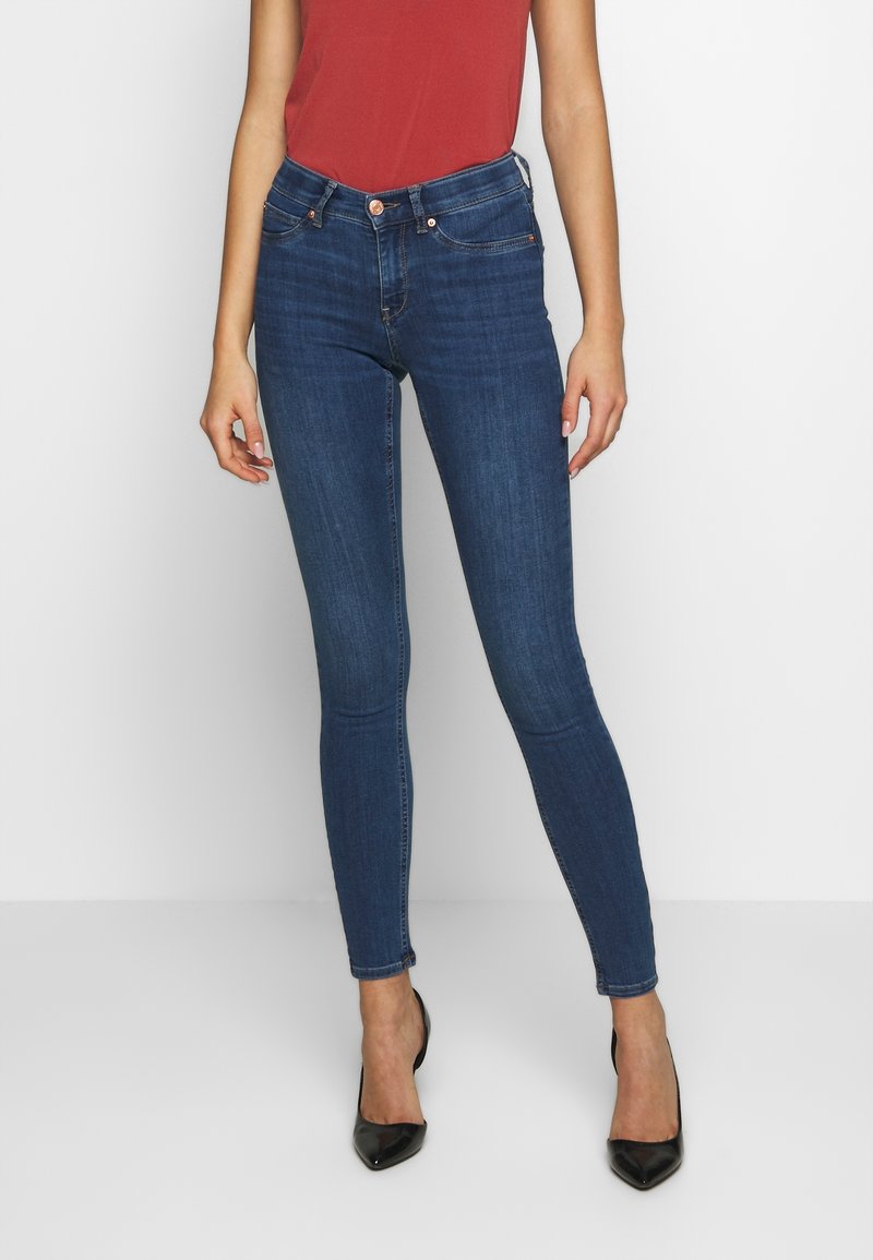 Jeans skinny blu in denim con vita alta, dotati di un bottone di rame, leggera scoloritura e cinque tasche. Indossati con tacchi neri.