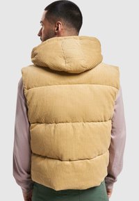 Chaleco de plumas de pana marrón con cuello alto y capucha incorporada. Presenta costuras verticales y secciones acolchadas para aislamiento.