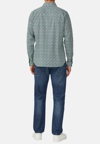 Chemise à manches longues verte à motifs floraux avec un col boutonné, portée avec un jean en denim bleu et des baskets blanches, vue de derrière.