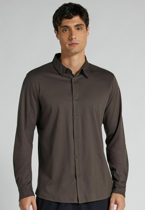 Mężczyzna w ciemnobrązowej koszuli na długi rękaw z guzikami, stojący przed jednolitym, jasnoszarym tłem, zwrócony przodem.