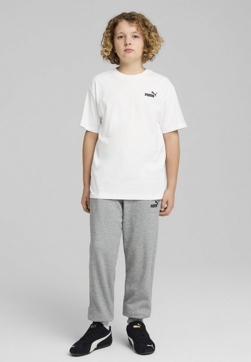 Biała bawełniana koszulka z czarnym logo Puma, w zestawie z szarymi dresami z polaru i czarnymi sneakersami. Model ma kręcone włosy.