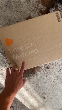 Brauner Karton mit orangefarbenem Logo und weißem Text "Liebe mich. Trage mich." Auf einer strukturierten Oberfläche positioniert. Hand berührt die Box.