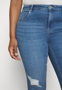 Blå denimjeans med nærbilde, som har detaljer med revne på kneet, fem lommer og glidelås med metallknapp.