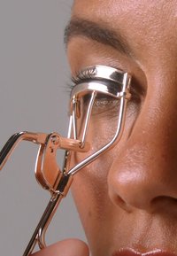 Rose gold eyelash curler designed with a metallic finish. Features curved arms and a rubber pad for grip, held against a closed eye.