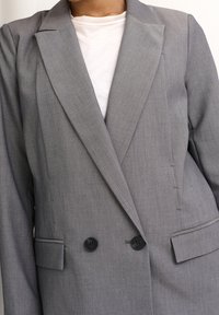 Grå blazer med dubbelknäppning, två svarta knappar, slagkrage och sidofickor. Slät textur, skräddarsydd passform, minimala detaljer.