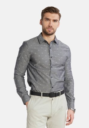 Camisa gris con estampado y pequeños logotipos en gris claro, con botones en la parte delantera y manga larga. Combinada con pantalones de color claro y un cinturón negro.