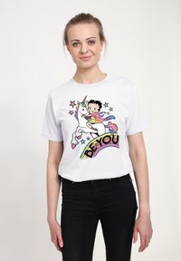 Bílý bavlněný tričko s potiskem kreslené dívky, která jezdí na jednorožci, obklopené barevnými hvězdami a textem "BUĎ SÁM SEBE" v duhových barvách.