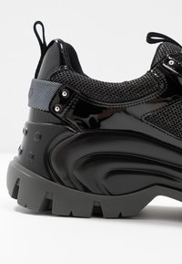 Černá sportovní bota s lesklým lakovaným povrchem, prodyšným síťovaným svrškem a masivní šedou podešví se hexagonálními vzory a akcenty.