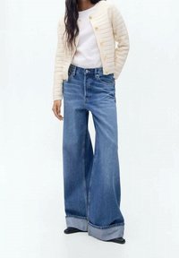 Jean en denim bleu à jambe large et taille haute, avec un design classique à cinq poches et fermeture à bouton. Complété par un cardigan texturé crème.