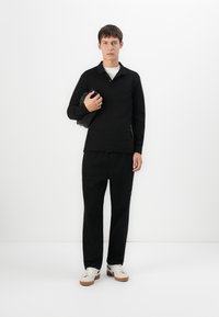 Tânăr îmbrăcat cu o cămașă neagră cu mâneci lungi și pantaloni negri, ținând un obiect negru pliat, stând în fața unui fundal alb simplu.