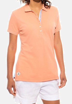 Kvinde iført en lakserød polo med korte ærmer og knapper samt hvide shorts, stående mod en ensartet lys baggrund.