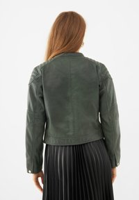 Tumma vihreä nahkatakki, jossa on rakenteelliset olkapäät, näkyvät saumat ja istuva leikkaus. Korostuu sileällä, matalla pinnalla.