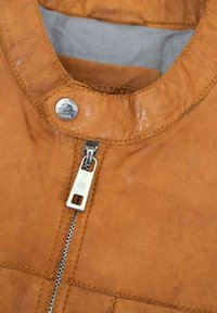 Tanfarbene Lederjacke mit rundem Halsausschnitt, silberner Hardware mit Reißverschluss und kontrastierenden Nähten. Weicher Stoff mit leichtem Glanz.