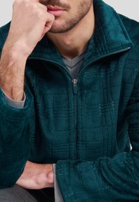 Chaqueta de forro polar en color verde azulado con un patrón de cuadrícula texturizado, cuello alto y cierre de cremallera. Capa interior gris visible. Material suave con un ajuste relajado.