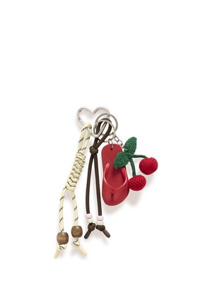 Frau hält eine beige Tragetasche mit schwarzen Sprenkeln und einem Kirsch-Schlüsselanhänger, trägt cremefarbene Hosen mit weitem Bein und braune offene Sandaletten.