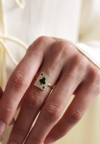 Anillo de oro con un diseño rectangular que incluye un trébol verde incrustado, rodeado de pequeñas piedras brillantes y dos pequeños acentos verdes.