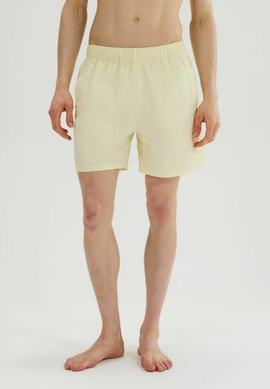 Hombre con pantalones cortos elásticos de cintura amarilla clara, de pie descalzo contra un fondo blanco liso.