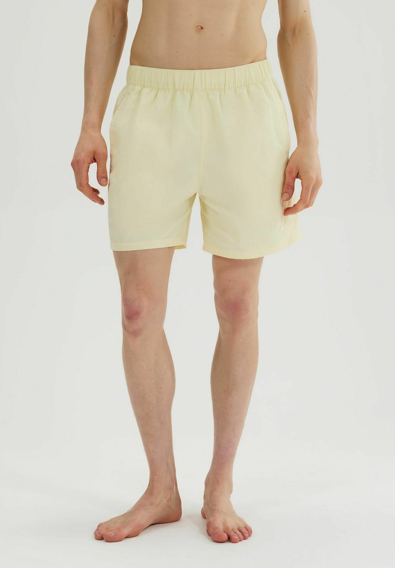 Άνδρας με ανοιχτό κίτρινο ελαστικό σορτς, όρθιος ξυπόλητος μπροστά σε απλό λευκό φόντο.