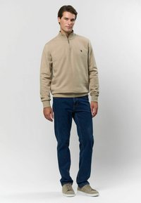 Béžová mikina so zipsom a dlhými rukávmi, skombinovaná s modrými džínsami a svetlosivými teniskami. Minimalistické logo na hrudi.