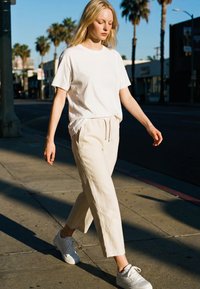 Blonde woman walking on sunlit sidewalk wearing white T-shirt, cream pants, and white sneakers with palm trees in background.
