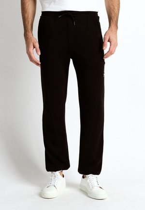 Mand iført sorte sweatpants med snøre og hvide sneakers, stående mod en ensfarvet baggrund, midtkropsvisning.
