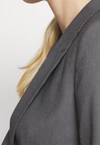 Blazer gris à fines rayures avec un col ajusté, une texture lisse et un éclat subtil. Présente une silhouette épurée et des détails de couture précis.