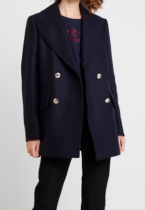 Femme portant un manteau bleu marine croisé à larges revers et boutons métalliques, sur un haut foncé et un pantalon noir, debout devant un fond clair.