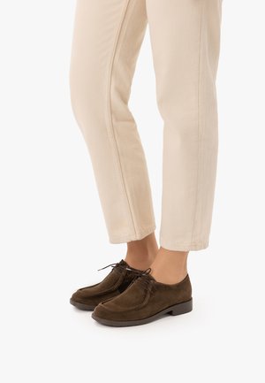 Person, die beige Hose und dunkelbraune Wildleder-Schnürschuhe trägt, steht auf einer weißen Oberfläche.