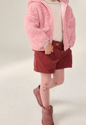 Enfant portant une veste rose duveteuse, un pull à col roulé blanc, un short en velours côtelé rouge, des collants roses à motifs de chats et des bottes roses, debout les jambes croisées.