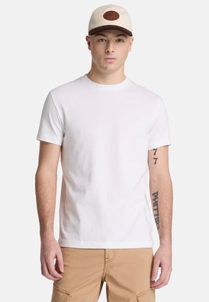 Giovane uomo con una t-shirt bianca a maniche corte, pantaloni cargo beige e un cappellino beige e marrone, in piedi davanti a uno sfondo semplice.