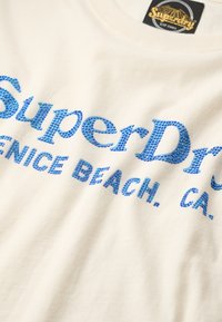 Hvid bomulds t-shirt med blå glitrende tekst, der siger "SuperDry Venice Beach, CA." Blød tekstur med rund hals og korte ærmer.