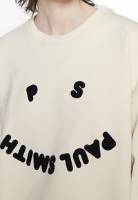 Beige mikina s černým, plyšovým nápisem "PAUL SMITH" a designem usměvavé tváře. Materiál vypadá měkký a texturovaný. Kulatý výstřih.
