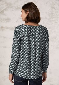 Pitkähihainen paita tummanvihreällä valkoisella houndstooth-kuviolla. Pehmeä kangas, jossa on pieni kiilto, pyöreä pääntie ja poimutuskuvio.
