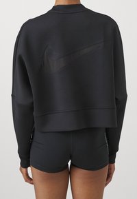 Svart cropped sweatshirt med ribbad halsringning, med en tonad Nike-logotyp på baksidan. Matchas med svarta tighta shorts. Slät textur.