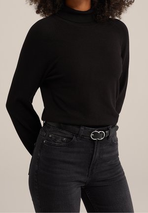 Kobieta ubrana w czarny, długi rękaw golf wsunięty w wysokowalne czarne dżinsy z czarnym paskiem z srebrną klamrą.