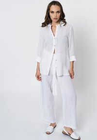 Witte linnen shirt en bijpassende brede broek, met een knoopsluiting en opgerolde mouwen, gedragen met witte instappers. Soepele textuur.